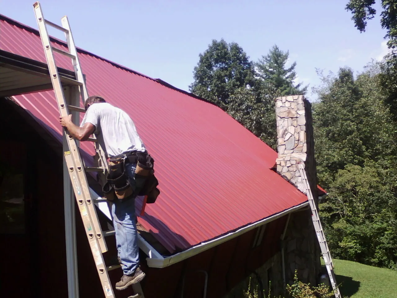 Roofer on a ladder beside a red metal roof and stone chimney — Soffit & Fascia Repair by Roofing Brothers in Wapello